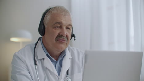 Un-Médico-Anciano-Vestido-Con-Bata-Blanca-Y-Estetoscopio-En-El-Cuello-Está-Consultando-A-Un-Paciente-En-Línea-Usando-Tecnología-De-Videollamada-En-Una-Computadora-Portátil